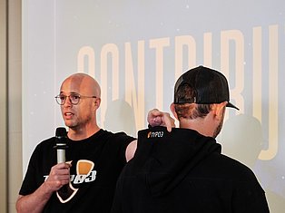 Two men speaking at a TYPO3 conference, one holding a microphone wearing glasses and a black shirt
