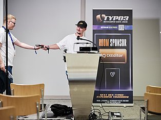 Two men at TYPO3 Developer Days 2020 conference, one speaking at podium with microphone, other holding microphone, with sponsor banner visible