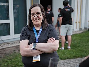 Woman with glasses and ID badge smiling with arms crossed at outdoor event