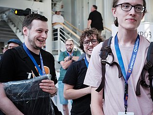 Group of smiling people wearing blue lanyards at an indoor event or conference, holding materials and standing together.