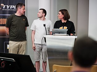 Three developers presenting at TYPO3 Developer Days conference with laptop and microphone on stage