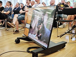 Group of people sitting in chairs watching a monitor displaying a dog's face during a presentation or demonstration event.