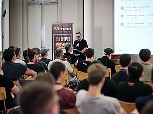 Speaker presenting at TYPO3 Developer Days conference to seated audience with sponsor banner and presentation slides visible