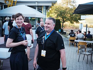 Two people wearing lanyards at an outdoor tech conference event with attendees and tents in background