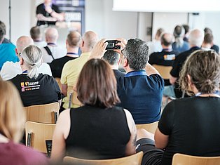 Man in blue shirt taking photo with smartphone at crowded indoor event or conference with seated audience