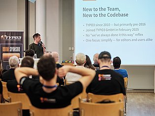 Speaker presenting at TYPO3 conference about joining team and codebase, with audience seated in auditorium