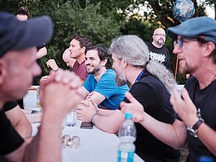 Group of men gathered outdoors around a table, smiling and socializing at an outdoor event with green foliage in background.