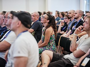 Diverse audience seated at conference, woman in colorful patterned dress and glasses attentively listening among attendees wearing lanyards