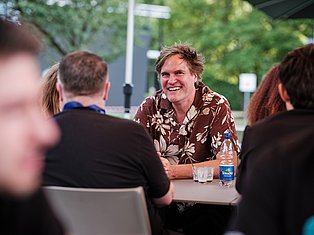 Man in brown floral shirt smiling while conversing with people at an outdoor table with water bottles
