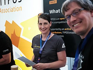 Woman wearing TYPO3 conference badge smiling at event with yellow branding backdrop and other attendees