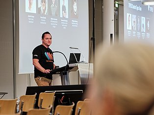 Man presenting at podium with team member photos displayed on projection screen behind him