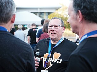 Man wearing glasses and lanyard badge smiling while holding a beer bottle at an outdoor event with other people