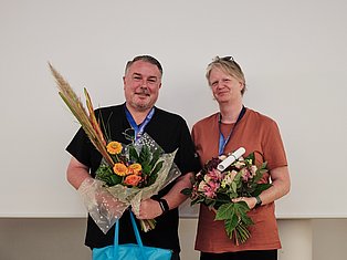 Two people wearing lanyards holding flower bouquets and a certificate against a white background