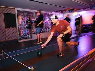 Man in red shirt bowling in neon-lit indoor venue with other people and colorful purple and orange lighting