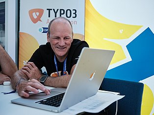 Man at TYPO3 conference smiling while using tablet and laptop at desk