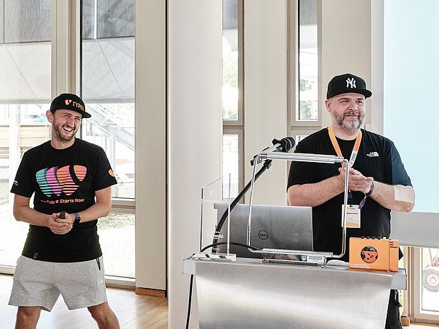 Two speakers at TYPO3 Developer Days standing at the front of a room, one smiling in a TYPO3 T-shirt and cap, the other applauding beside a podium with a laptop and microphone.