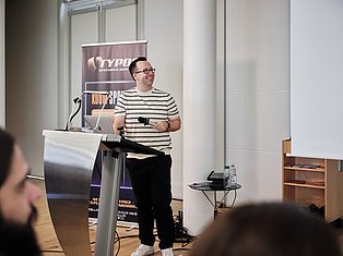 Man in striped shirt speaking at podium with microphone at tech conference with TYPO3 banner