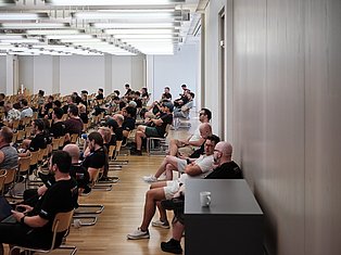Large lecture hall with many students seated in wooden chairs during a presentation or class session