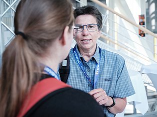 Man wearing glasses and blue lanyard badge speaking with woman in industrial warehouse setting