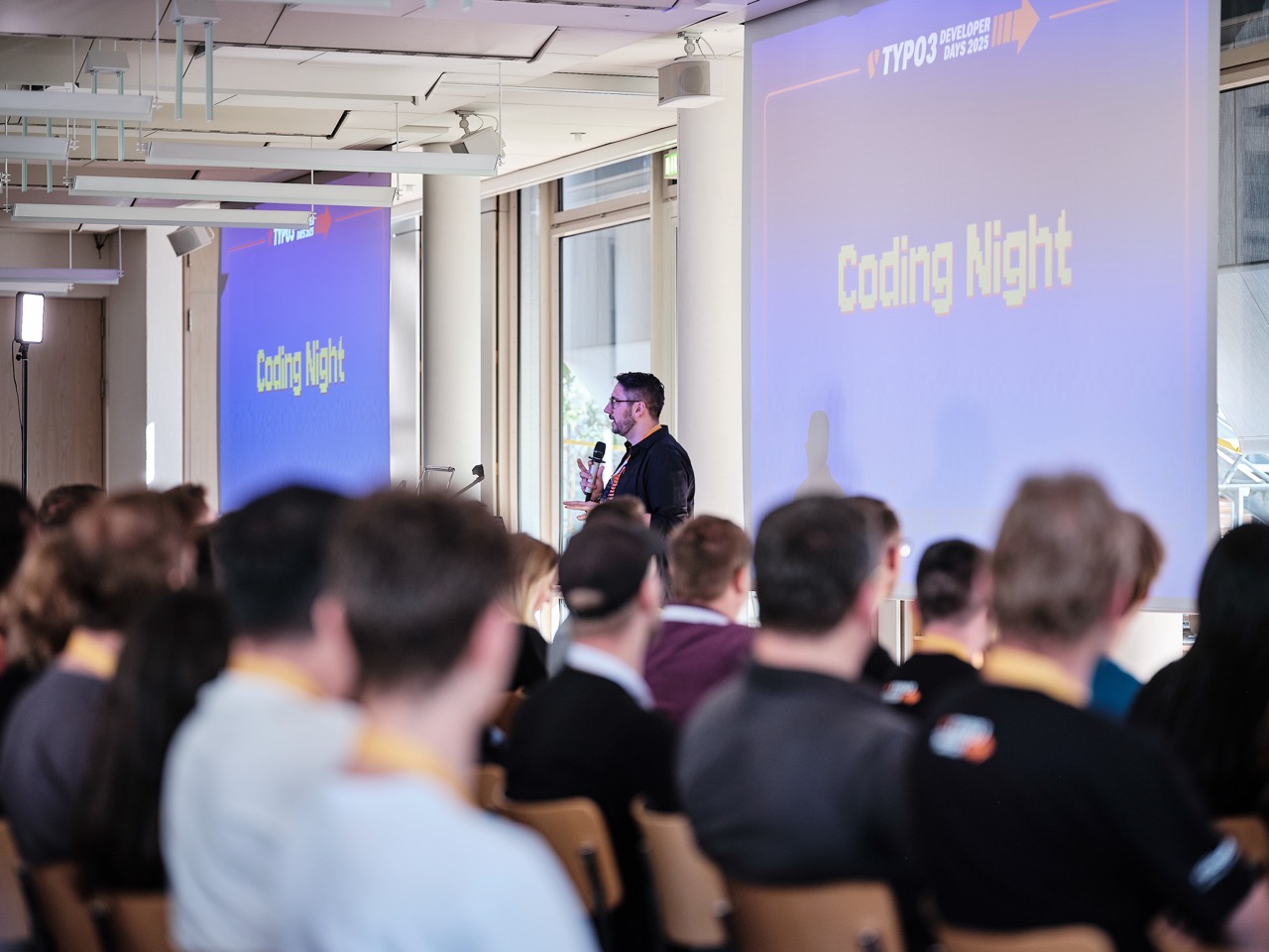 Speaker holding a microphone in front of a screen reading ‘Coding Night’ at TYPO3 Developer Days, with an audience seated in the foreground.