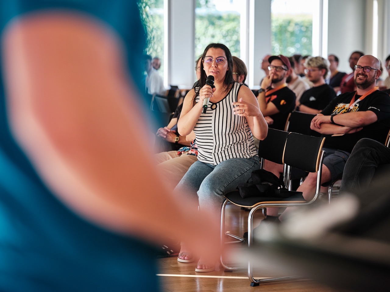 A person in the audience holding a microphone and asking a question during an event; other attendees sit in the background.